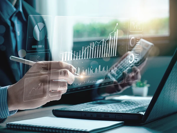 Eine Person in einem Anzug nutzt einen Stift, um auf ein digitales Diagramm mit Grafiken und Statistiken zu zeigen, während sie an einem Laptop arbeitet. Im Hintergrund sind Pflanzen und Fenster zu sehen, die Tageslicht hereinlassen.