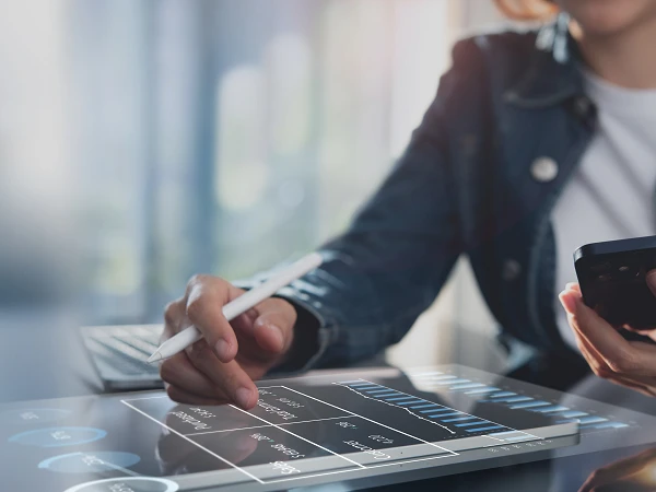 Eine Person in einer Jacke nutzt ein Smartphone in der rechten Hand, während sie mit einem Stylus auf einem Tablet mit grafischen Daten und Diagrammen arbeitet. Im Hintergrund ist ein Laptop sichtbar.