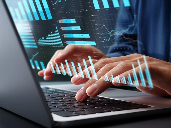 Zwei Hände tippen auf der Tastatur eines Laptops, während grafische Darstellungen von Datenanalysen, wie Balken- und Liniendiagramme, über den Bildschirm projiziert werden.