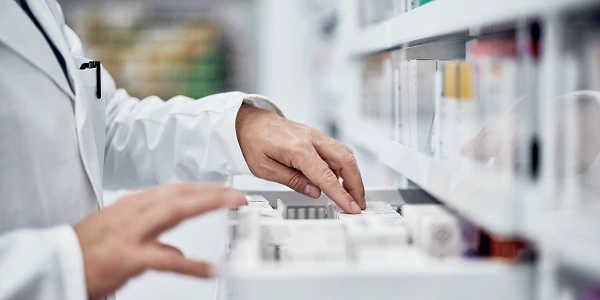 Ein Apotheker in weißem Kittel greift mit der linken Hand nach einem Medikamentenbehälter aus einem Regalsystem, während die rechte Hand auf der Ablage ruhen bleibt, um eine sorgfältige Auswahl zu treffen.