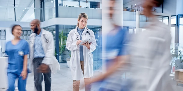 Eine Ärztin in weißem Kittel steht mit einem Tablet in der Hand in einem modernen Krankenhaus, während unscharfe Personen in blauer und weißer Berufsbekleidung im Hintergrund vorbeigehen.