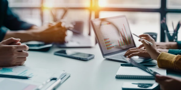Mehrere Personen sitzen an einem Tisch, nutzen Laptops und Tablets, während sie Diagramme und Grafiken auf dem Bildschirm analysieren. Auf dem Tisch liegen Notizblöcke, Stifte und Smartphones. Tageslicht strömt durch große Fenster.
