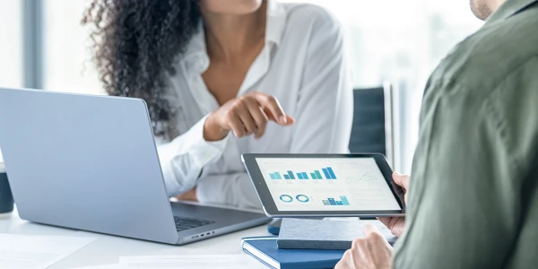 Eine Person mit lockigem Haar zeigt auf eine Grafik auf einem Tablet, während eine andere Person am Tisch sitzt und einen Laptop vor sich hat, in einem modernen Büro.