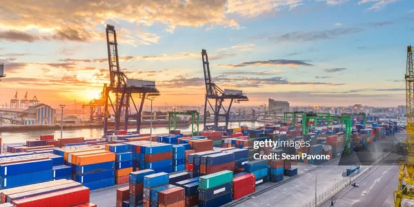 Hafen mit mehreren großen Kränen, die Container bewegen, während die Sonne am Horizont aufgeht und den Himmel in warmen Farben erleuchtet. Containerstapel sind ordentlich auf dem Boden angeordnet.