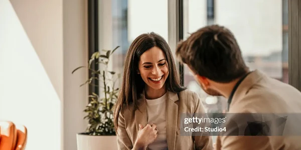 Eine Frau mit langen, dunklen Haaren sitzt in einem modernen Innenraum und lächelt, während sie mit einem Mann spricht, der ihr gegenüber sitzt und eine Mappe in der Hand hält. Im Hintergrund ist eine Pflanze zu sehen und große Fenster lassen Tageslicht herein.