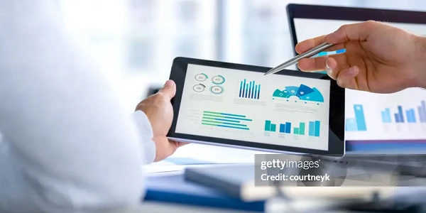 Ein Grafikanalyst zeigt mit einem Stift auf ein Tablet, das verschiedene Diagramme und Datenvisualisierungen anzeigt, während eine andere Person daneben sitzt und zuhört. Im Hintergrund sind weitere Diagramme auf einem Computerbildschirm zu sehen.