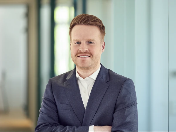 Ein Mann mit rotem Haar und Schnurrbart trägt einen dunkelblauen Anzug und steht mit verschränkten Armen vor einer hellen, modern gestalteten Büroeinrichtung.