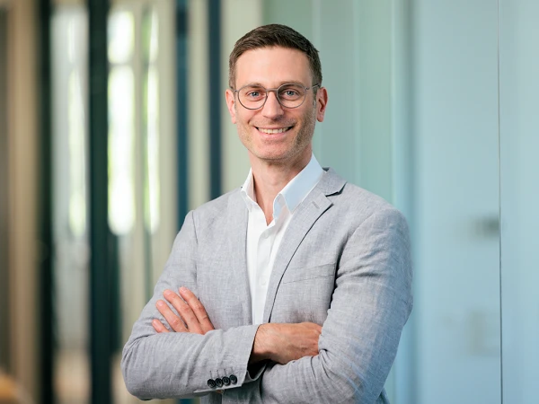 Ein Mann mit Brille trägt einen grauen Blazer und steht lächelnd mit verschränkten Armen vor einem modernen, lichtdurchfluteten Büro.