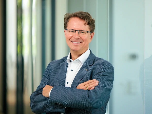 Ein Mann mit Brille und einem lächelnden Gesicht steht mit verschränkten Armen in einem modernen Büroumfeld. Er trägt einen blauen Blazer und ein weißes Hemd mit Knöpfen. Im Hintergrund sind verschwommene Wände und Fenster zu erkennen.