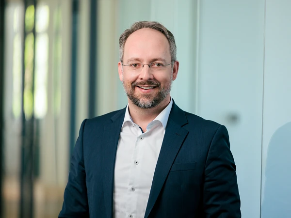 Ein lächelnder Mann in einem dunkelblauen Anzug mit Brille steht in einem modernen Büroraum, umgeben von großen Fenstern und hellen Wänden.