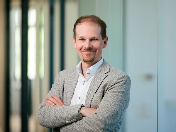 Ein Mann mit kurzen Haaren und einem gepflegten Bart steht in einem modernen Büro, lächelt und hat die Arme vor der Brust verschränkt. Er trägt einen hellen Blazer über einem weißen Hemd. Hinter ihm sind unscharfe Büroelemente zu erkennen.