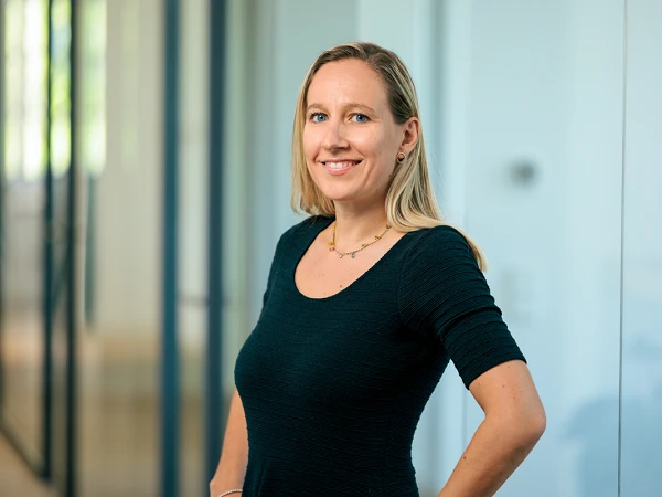 Eine Frau mit langen blonden Haaren trägt ein schwarzes, enges Oberteil und steht lächelnd in einem modernen Büro mit großen Fenstern.