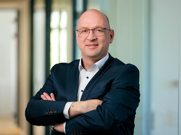 Ein Mann mit Brille steht in einem modernen Büro mit verschränkten Armen, trägt einen dunkelblauen Anzug und lächelt in die Kamera.