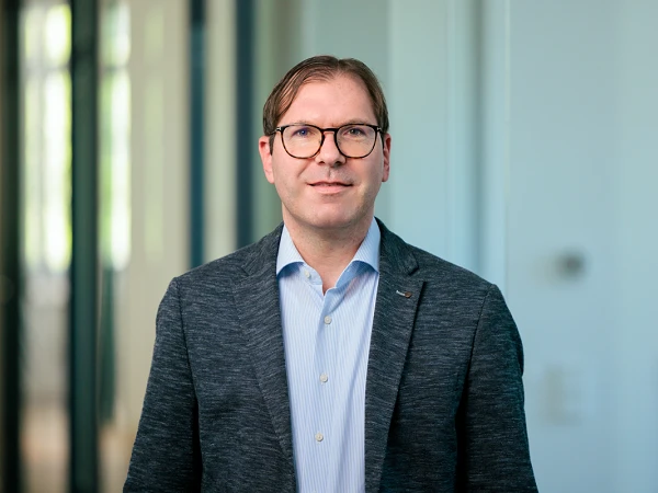 Ein Mann mit Brille, wearing a dunkle Jacke über einem hellen Hemd, steht in einem modernen Büro mit großen Fenstern im Hintergrund.