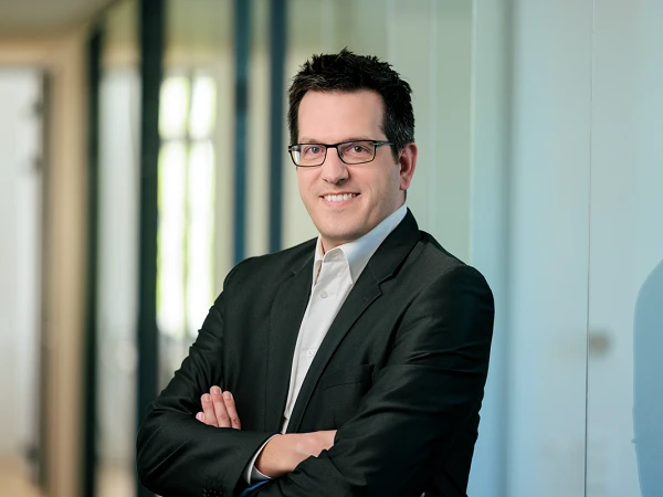 Ein Mann in einem schwarzen Anzug mit einem weißen Hemd und einer dunklen Brille steht in einem modernen Bürogebäude, lächelt und hat die Arme verschränkt. Im Hintergrund sind Glaswände und weiches Licht zu sehen.
