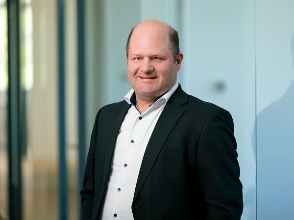 Ein Mann in einem schwarzen Anzug mit einem weißen Hemd steht lächelnd vor einer Glaswand in einem modernen Bürogebäude.