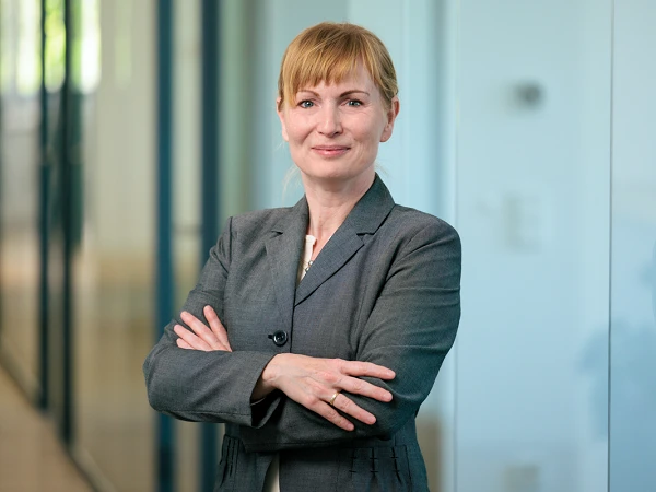 Eine Geschäftsfrau mit einem selbstbewussten Lächeln steht mit verschränkten Armen vor einer Glaswand in einem modernen Bürogebäude. Sie trägt einen grauen Anzug und hat blondes, schulterlanges Haar.