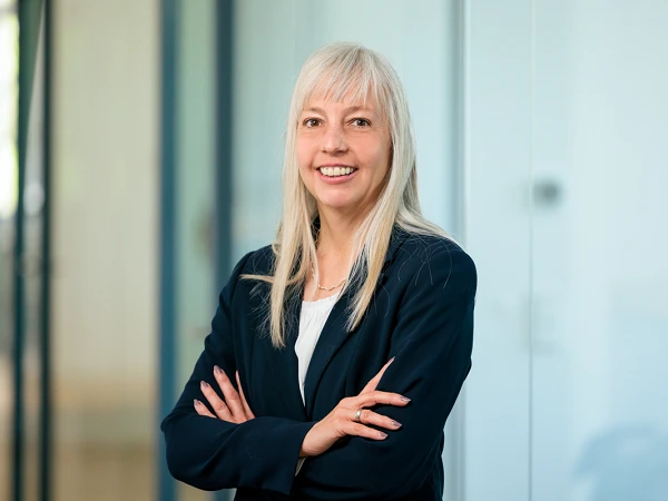Eine Frau mit langen, blonden Haaren steht mit verschränkten Armen und lächelt in einem modernen Büroumfeld. Sie trägt einen dunklen Blazer und hat einen professionellen, freundlichen Ausdruck.