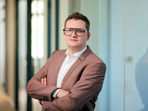 Ein junger Mann mit Brille in einem eleganten braunen Anzug steht mit verschränkten Armen in einem modernen Büro. Er wirkt selbstbewusst und schaut direkt in die Kamera.