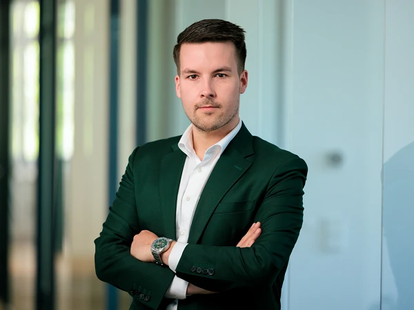 Ein junger Mann in einem dunklen Anzug mit einem weißen Hemd steht in einem modernen Büro, mit verschränkten Armen und einem selbstbewussten Ausdruck.
