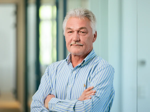 Ein älterer Mann mit grau meliertem Haar und einem Schnurrbart steht mit verschränkten Armen in einem modernen Büroflur. Er trägt ein gestreiftes Hemd und schaut direkt in die Kamera. Die Wände im Hintergrund sind hell und geben einen klaren, professionellen Eindruck.