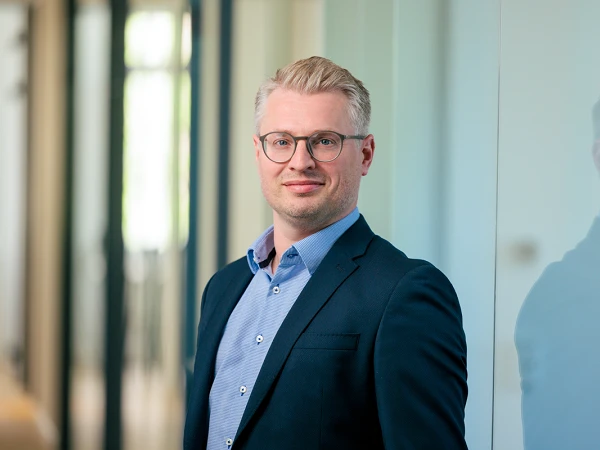 Ein Mann mit Brille und kurzem, blonden Haar steht in einem modernen Büro. Er trägt ein dunkles Sakko und ein hellblaues Hemd und lächelt leicht in die Kamera. Im Hintergrund sind unscharfe Büroelemente zu sehen.
