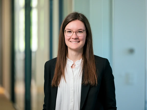 Eine Person mit langen, braunen Haaren und Brille steht vor einer hellen, modernen Büroumgebung und lächelt freundlich in die Kamera. Sie trägt ein schwarzes Blazer und eine weiße Bluse.