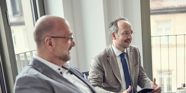 Zwei Männer sitzen an einem Tisch in einem hellen, modernen Büroraum; der Mann rechts spricht und hält ein Tablet in der Hand, während der Mann links aufmerksam zuhört.