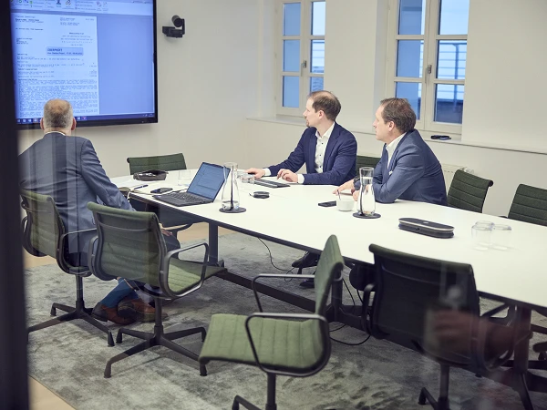 Drei Personen sitzen an einem Konferenztisch in einem hellen Besprechungsraum und schauen auf einen großen Bildschirm, auf dem ein Dokument angezeigt wird. Zwei der Personen sind in Anzügen gekleidet, während eine Person von hinten sichtbar ist und in einem karierten Anzug sitzt. Auf dem Tisch stehen Laptops und Wasserkrüge.