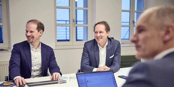 Zwei Männer in formeller Kleidung sitzen an einem Tisch in einem Besprechungsraum und lächeln, während ein dritter Mann in einem Anzug von der Seite auf einen Laptop schaut. Auf dem Tisch stehen Gläser mit Wasser und eine Tasse, während auf dem Laptop ein Dokument sichtbar ist. Durch die Fenster scheint Tageslicht.