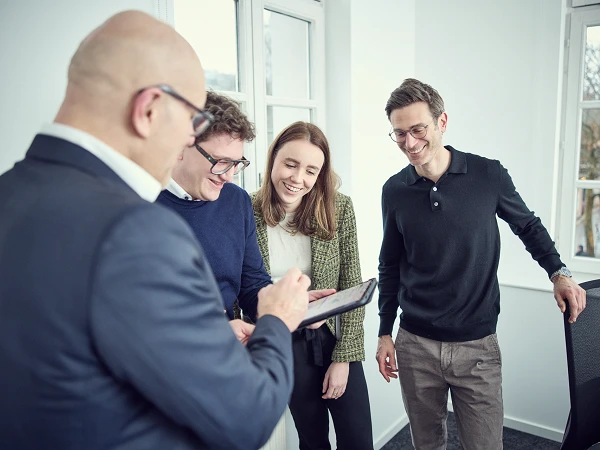 Eine Gruppe von vier Personen in einem hellen Büroraum schaut auf ein Tablet, während sie miteinander sprechen und lächeln.