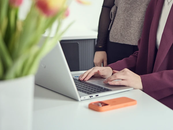 Zwei Frauen stehen in einem modernen Büro und schauen lächelnd auf einen Laptop, während im Hintergrund eine Präsentation auf einem Bildschirm angezeigt wird. Auf dem Tisch steht eine Vase mit bunten Tulpen.