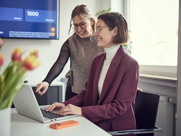 Zwei Frauen sitzen an einem Tisch in einem modernen Büro; eine von ihnen zeigt auf den Bildschirm, während die andere am Laptop arbeitet und lächelt. Auf dem Tisch liegen bunte Tulpen und ein Smartphone.