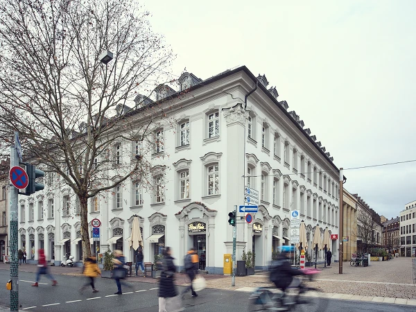 Ein elegantes, historisches Gebäude mit weißer Fassade und vielen Fenstern an einer Straßenecke, umgeben von Fußgängern und modernen Verkehrsschildern. Im Vordergrund sind unscharfe Passanten zu sehen, die die Straße überqueren, während die Bäume im Hintergrund verblasst sind.