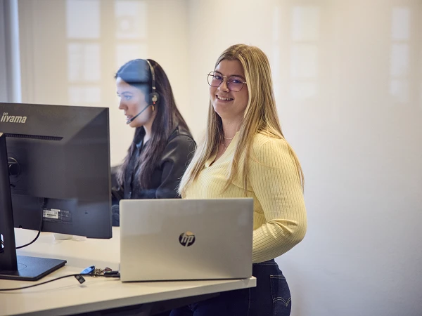 Eine lächelnde Frau mit langen blonden Haaren steht an einem modernen Schreibtisch, während sie in die Kamera schaut; neben ihr sitzt eine weitere Frau mit einem Headset am Computer.