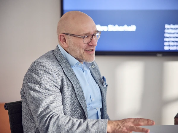 Ein Mann mit Brille und einem hellen Blazer lächelt und spricht engagiert, während er an einem Tisch sitzt. Im Hintergrund ist ein Bildschirm mit Text sichtbar.