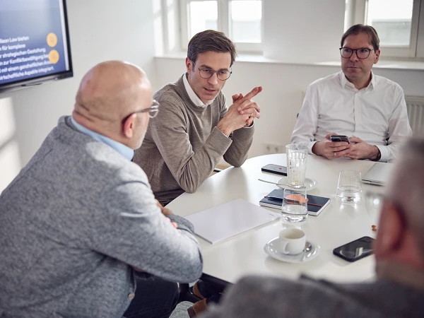 Ein Meeting mit mehreren Teilnehmern an einem Tisch. Ein Mann in einem grauen Pullover spricht anhand von Notizen, während zwei andere Männer ihm zuhören. Auf einem Bildschirm im Hintergrund sind Präsentationsinhalte zu sehen. Auf dem Tisch stehen Gläser und Tassen.