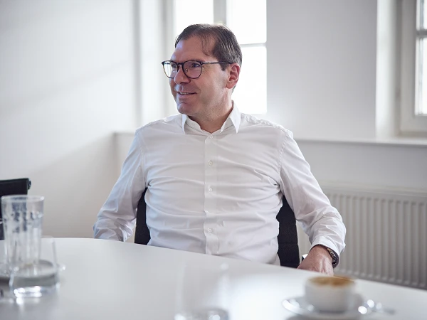 Ein Mann mit Brille sitzt an einem Tisch in einem modernen, hellen Raum und blickt entspannt zur Seite. Auf dem Tisch sind Gläser und eine Kaffeetasse zu sehen.