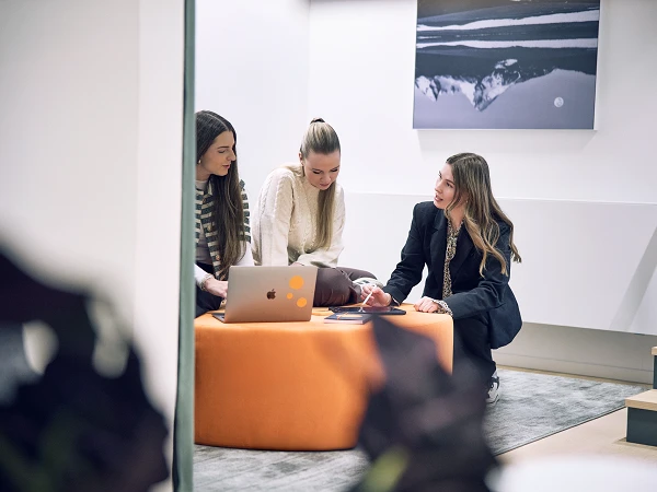 Drei Frauen sitzen gemeinsam auf einem orangen Pouf und schauen auf einen Laptop, während sie an einem Projekt arbeiten. Eine Frau hält ein Handy in der Hand und alle drei scheinen in ein Gespräch vertieft zu sein. Im Hintergrund ist ein abstraktes Bild an der Wand zu sehen.