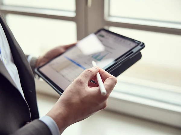 Ein Mann in einem Anzug hält ein Tablet in der Hand und verwendet einen Stylus, während er Daten und Diagramme auf dem Bildschirm betrachtet. Im Hintergrund sind Fenster zu sehen, die Tageslicht hereinlassen.