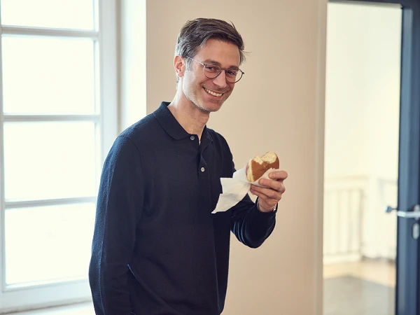Ein Mann mit Brille hält ein belegtes Brötchen in der Hand und lächelt freundlich, während er in einem hellen Raum steht.