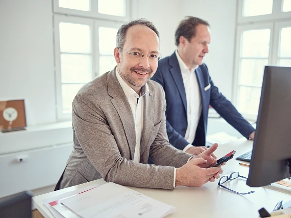 Zwei Männer in einem modernen Büro stehen an einem Tisch; einer von ihnen hält ein Smartphone in der Hand und lächelt in die Kamera, während der andere hinter einem Computerbildschirm steht und auf den Bildschirm schaut. Auf dem Tisch liegen einige Dokumente.