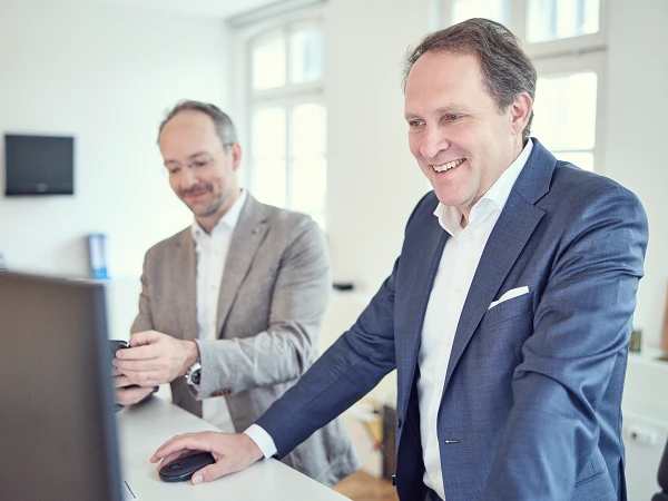 Zwei Männer in formeller Kleidung stehen an einem weißen Schreibtisch; einer von ihnen lächelt und schaut auf einen Computerbildschirm, während der andere mit einem Smartphone in der Hand beschäftigt ist. Im Hintergrund sind Büroelemente wie ein Fernseher und eine Uhr sichtbar.