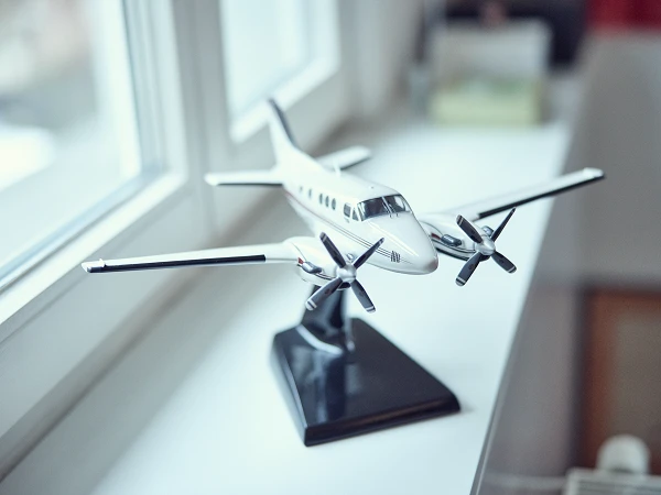 Ein Modellflugzeug mit zwei Propellern steht auf einem Fensterbrett und wird von Tageslicht beleuchtet. Im Hintergrund sind unscharf Möbel und eine Fensterbank zu sehen.