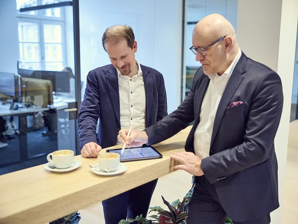 Zwei Männer stehen an einem Holztisch in einem modernen Büro, während einer von ihnen auf einem Tablet schreibt. Vor ihnen stehen zwei Tassen Kaffee. Der Raum ist hell und minimalistisch gestaltet, mit Büroarbeiten im Hintergrund.