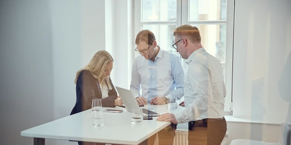 Drei Personen, zwei Männer und eine Frau, stehen an einem Tisch in einem modernen Büro und betrachten einen Laptop; auf dem Tisch stehen Wassergläser.