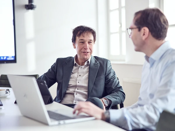 Zwei Männer sitzen an einem Tisch in einem modernen Besprechungsraum und führen ein Gespräch, während ein Laptop und Tassen auf dem Tisch stehen.