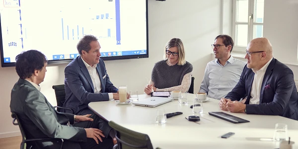 Eine Gruppe von fünf Personen sitzt an einem Konferenztisch in einem modernen Besprechungsraum. Auf einem großen Bildschirm im Hintergrund sind Diagramme und Grafiken zu sehen. Die Teilnehmer sind in Diskussion vertieft, während Notizen gemacht werden.
