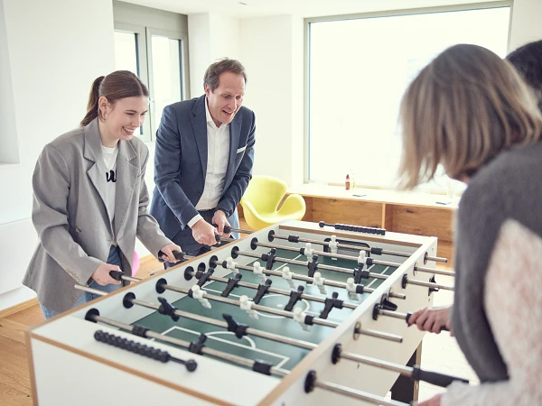 Zwei Personen stehen an einem Tischfußballspiel und spielen gegeneinander, während eine zweite Gruppe zuschaut. Der Mann trägt einen Anzug, die Frau eine große Bluse, und beide scheinen viel Spaß zu haben. Im Hintergrund sind helle Räume mit Fenstern und modernen Möbeln sichtbar.
