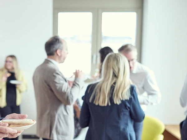 Eine Gruppe von Personen steht in einem hellen Raum, einige halten Teller mit Snacks, während andere miteinander sprechen. Im Vordergrund ist eine Person mit blonden Haaren und einem dunkelblauen Anzug zu sehen, die von hinten gezeigt wird. Im Hintergrund sind weitere Gäste erkennbar.